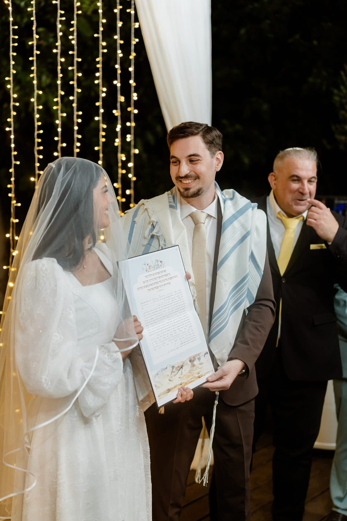 כלה וחתן עם טלית ומסמך מעוצב בידיים, מחייכים