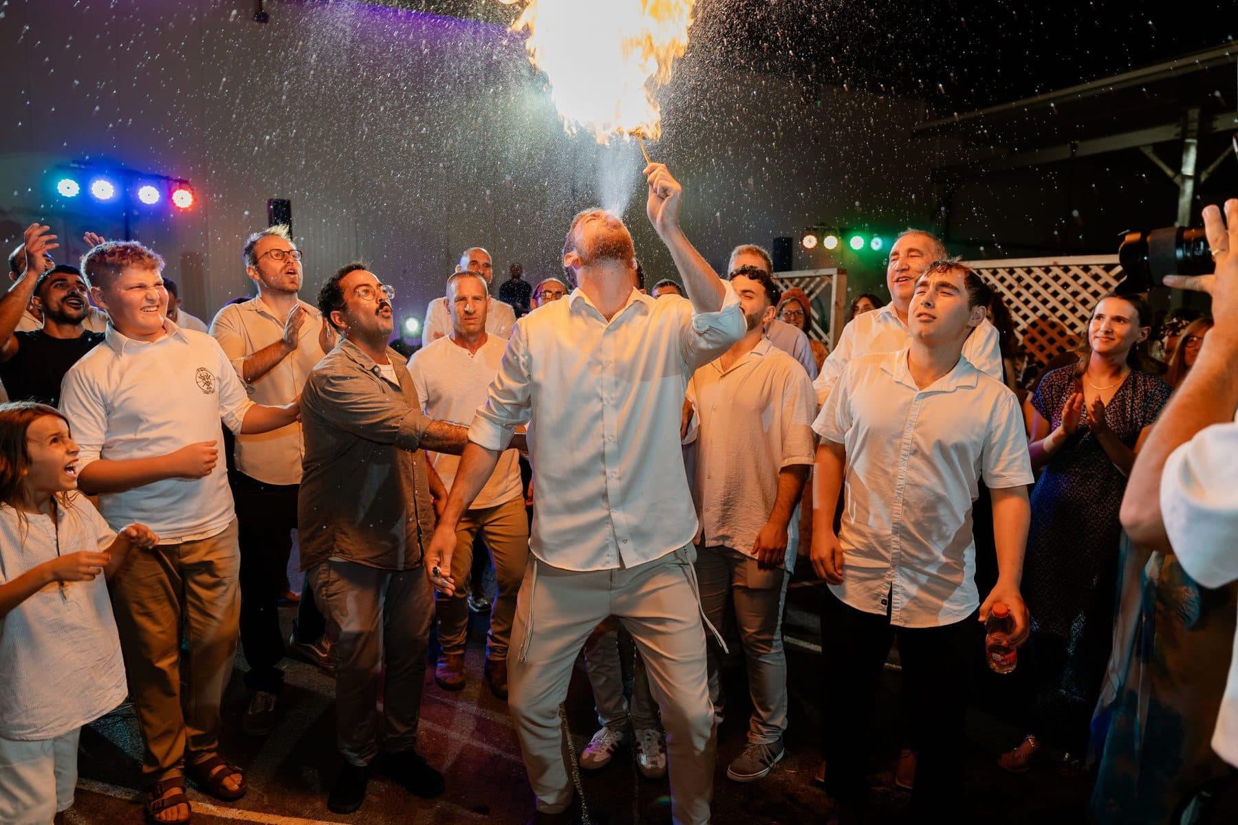 גברים רוקדים ברחבה בלילה, עם אורות צבעוניים ושפריצי אש